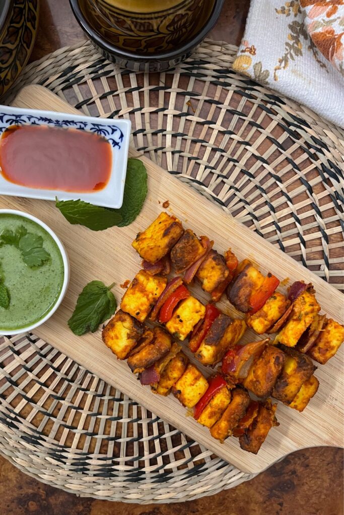 Paneer Tikka presented on a wooden board with a couple mint leaves served with green and red chutneys