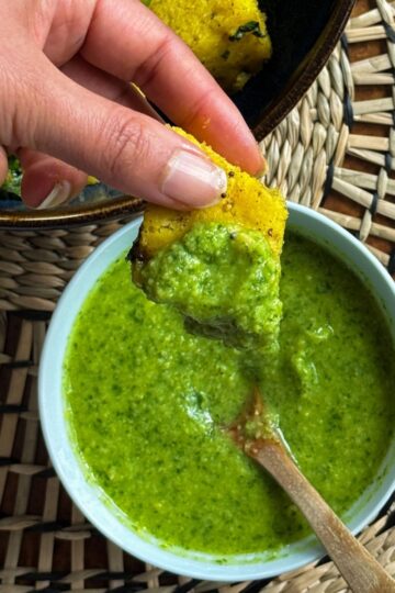 Dipping Khaman piece in a bowl of khaman chutney with a wooden spoon in it.