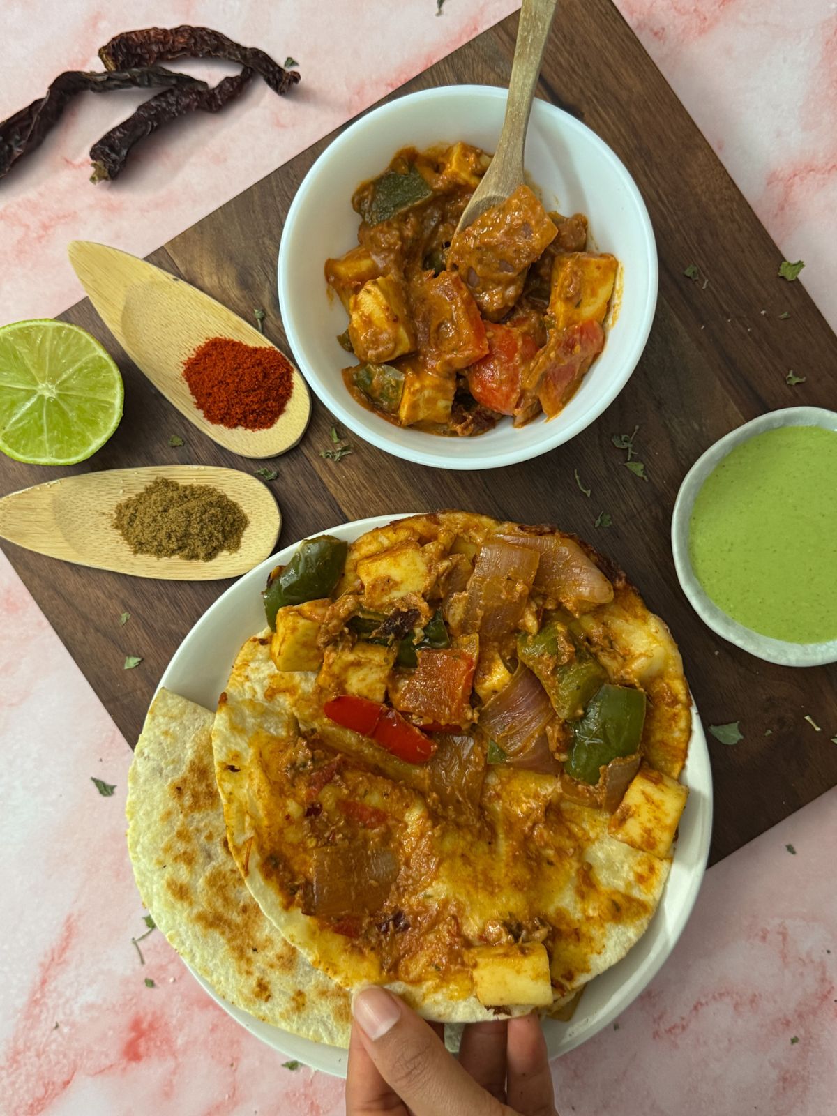 A deconstructed view of a tortilla showing the tikka filling into it presented with a green chutney, lime, spices and a white bowl filled with the tikka taco filling. 