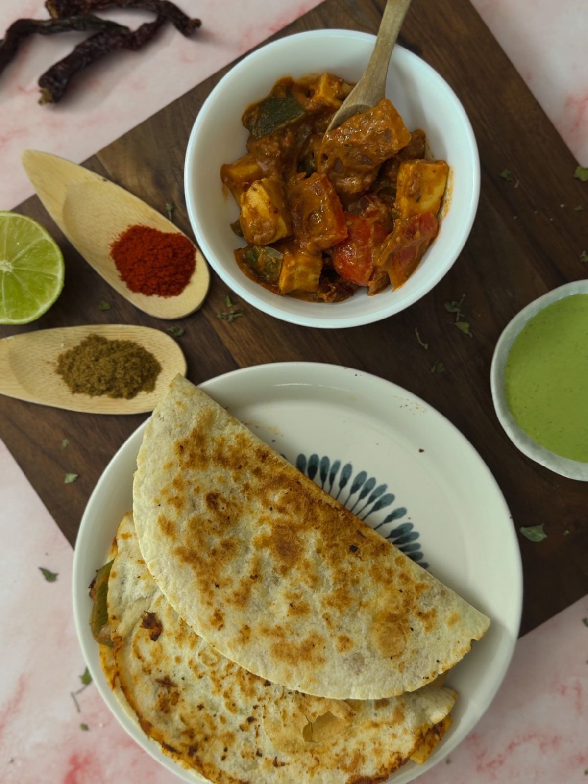 A plate with two tortillas presented with a green chutney, lime, spices and a white bowl filled with the tikka taco filling.
