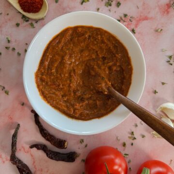 A white bowl with homemade tikka paste and a wooden spoon into it, surrounded by tomatoes, garlic and dried chillies and sprinkled kasoori methi