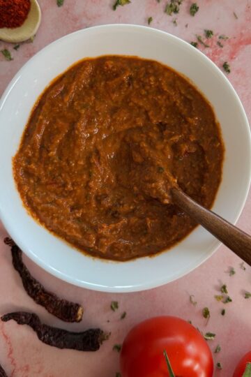 A white bowl with homemade tikka paste and a wooden spoon into it, surrounded by tomatoes, garlic and dried chillies and sprinkled kasoori methi