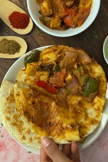 A deconstructed view of a tortilla showing the tikka filling into it presented with a green chutney, lime, spices and a white bowl filled with the tikka taco filling.