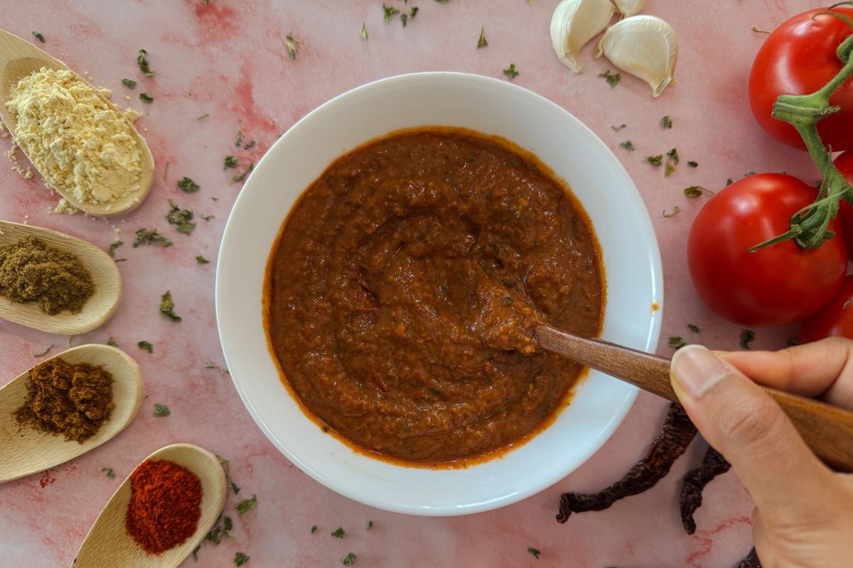 A white bowl with homemade tikka paste and a wooden spoon with my fingers holding the spoon, surrounded by tomatoes, garlic and spice spoons.